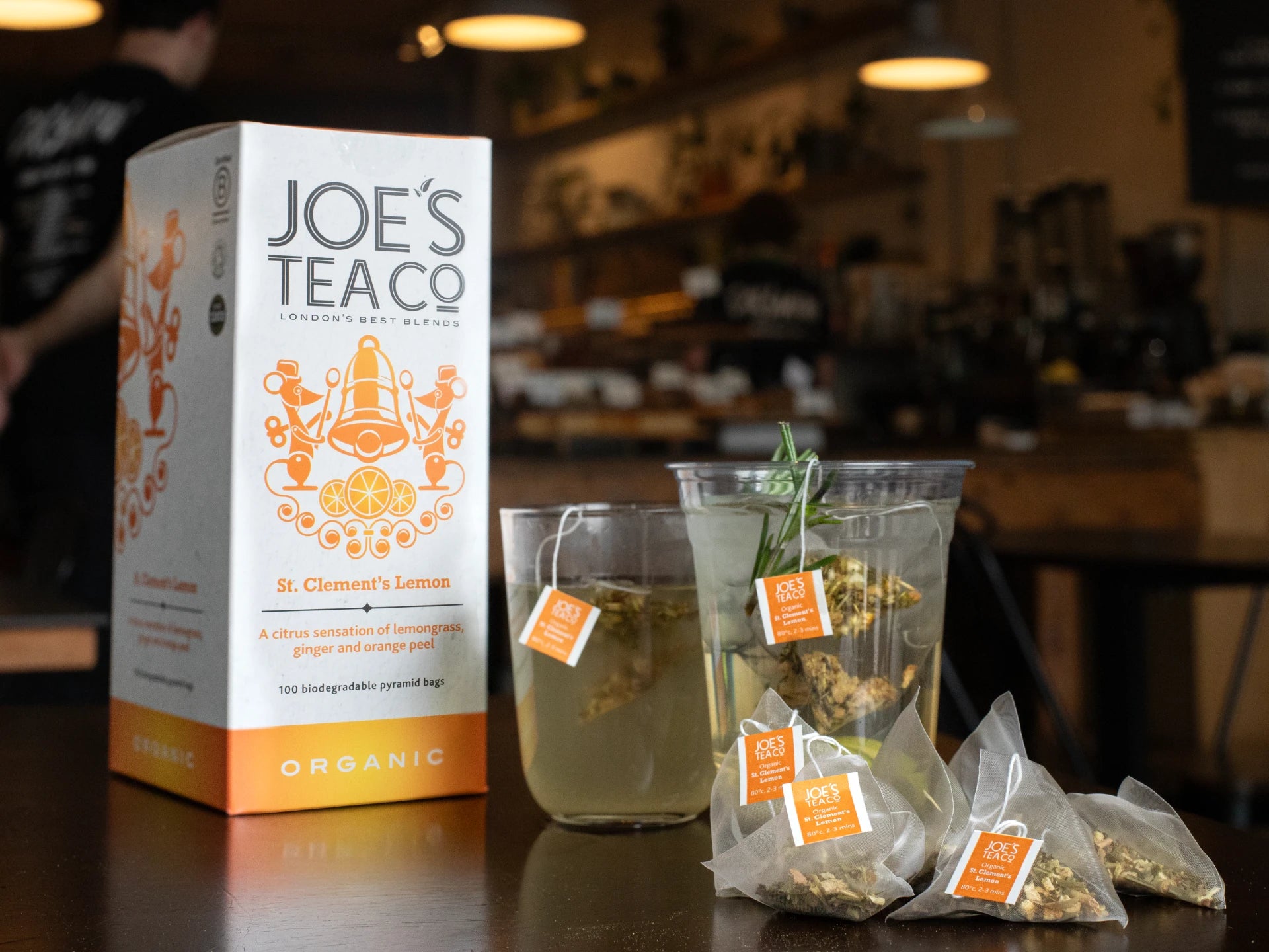 Joe's Tea Co - St. Clements Lemon packaging with two cups of tea and tea bags on a table in a bar setting.