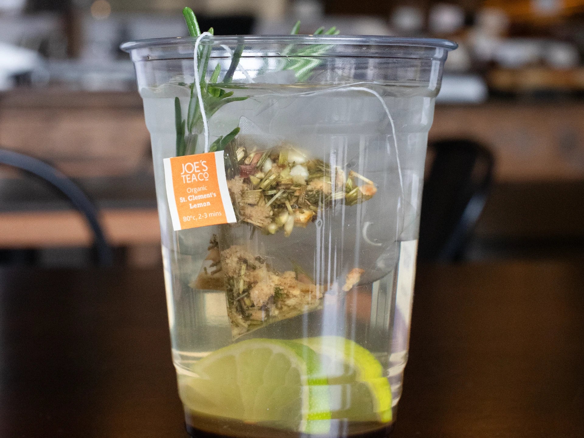Clear plastic cup with Joe's Tea co  iced tea, lime, and rosemary on a wooden table.