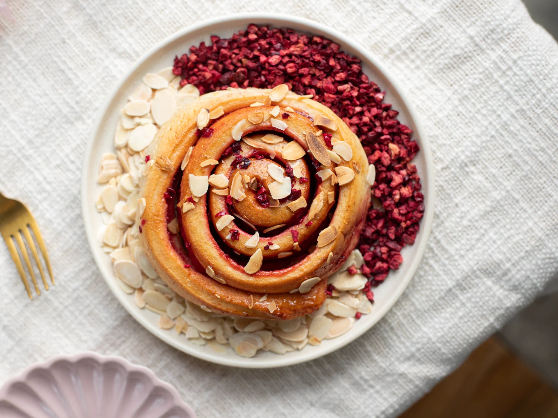 Sticky Cherry Bakewell Bun with almonds and dried berries on a white plate with a gold fork.
