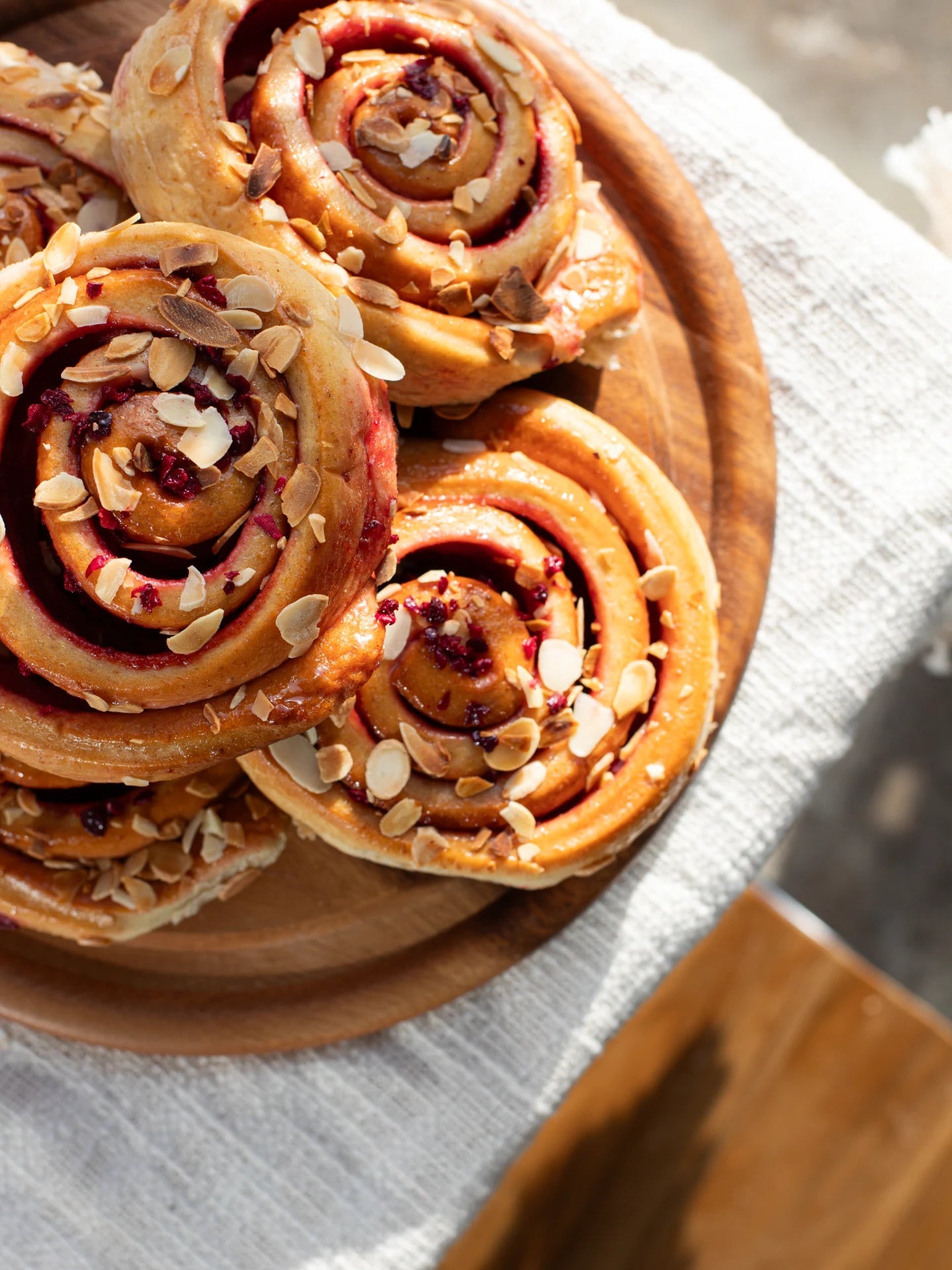 Wooden plate with Sticky Cherry Bakewell Bun topped with nuts on a light fabric background