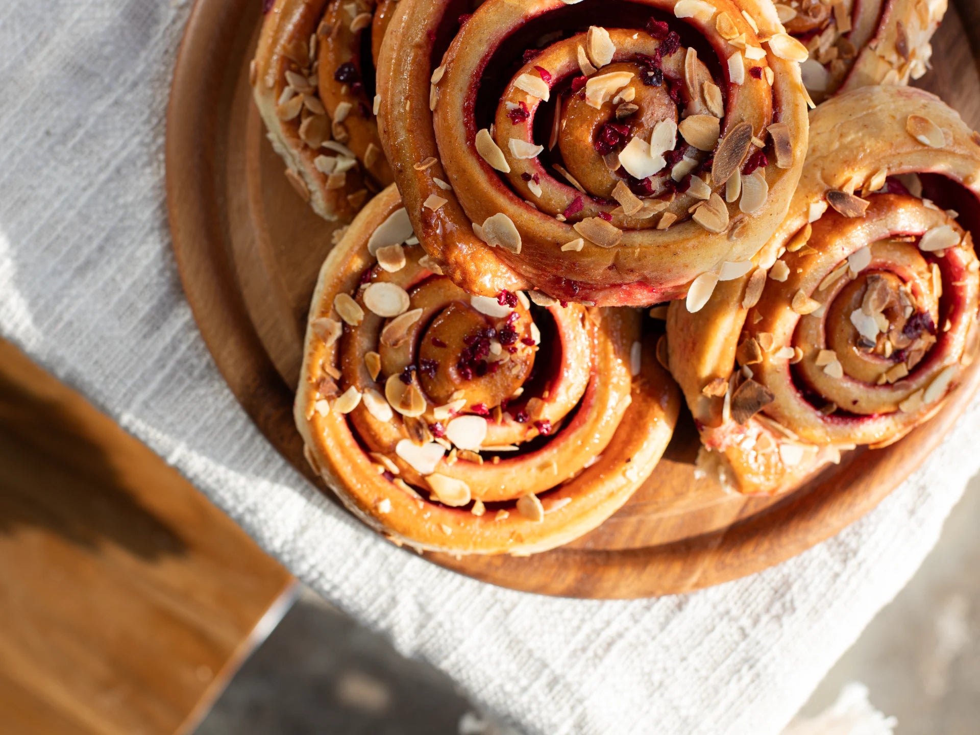 Wooden plate with Sticky Cherry Bakewell Bun topped with nuts on a light fabric background