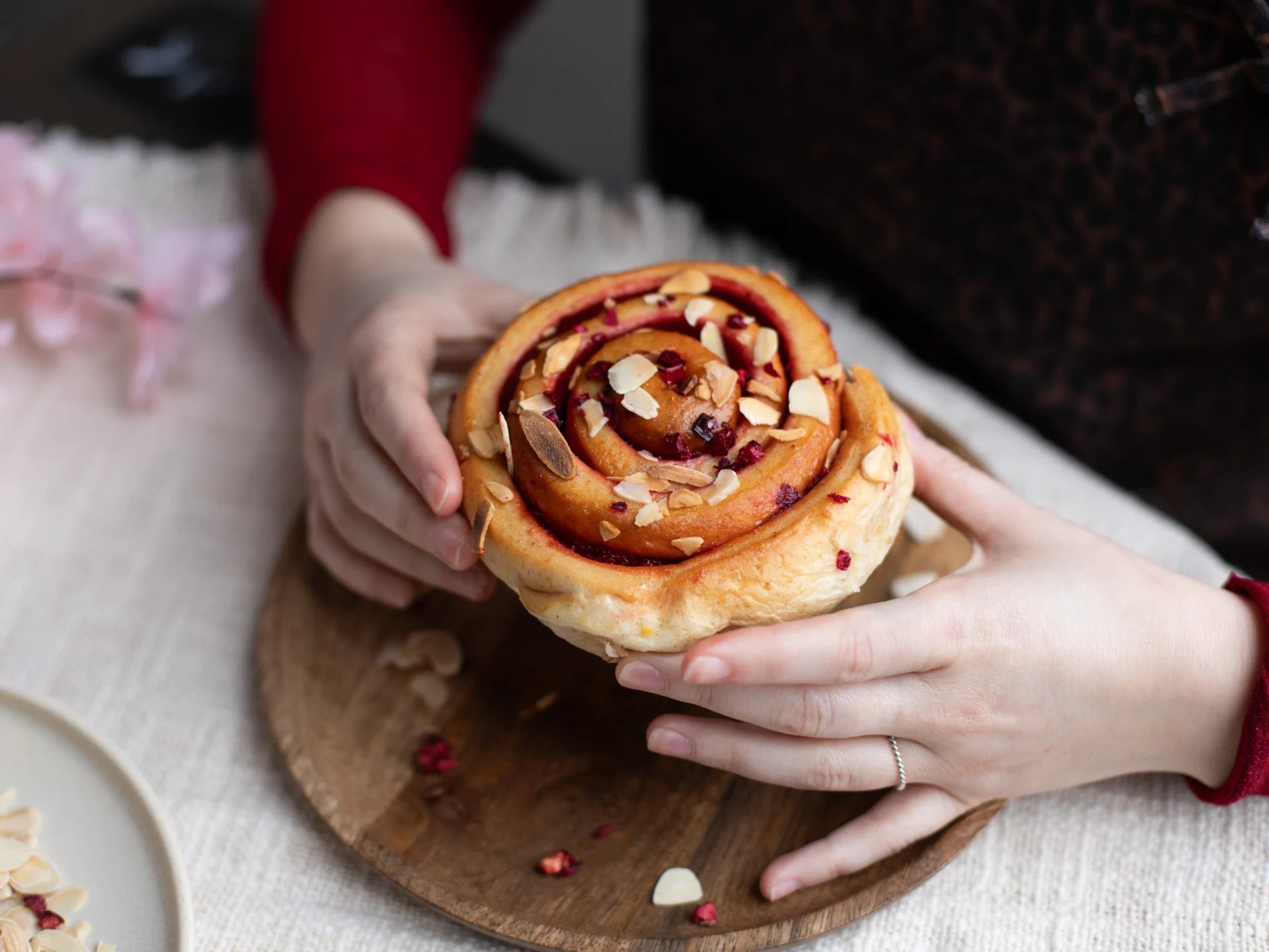 Hands holding a Sticky Cherry Bakewell Bun with toppings on a wooden board.