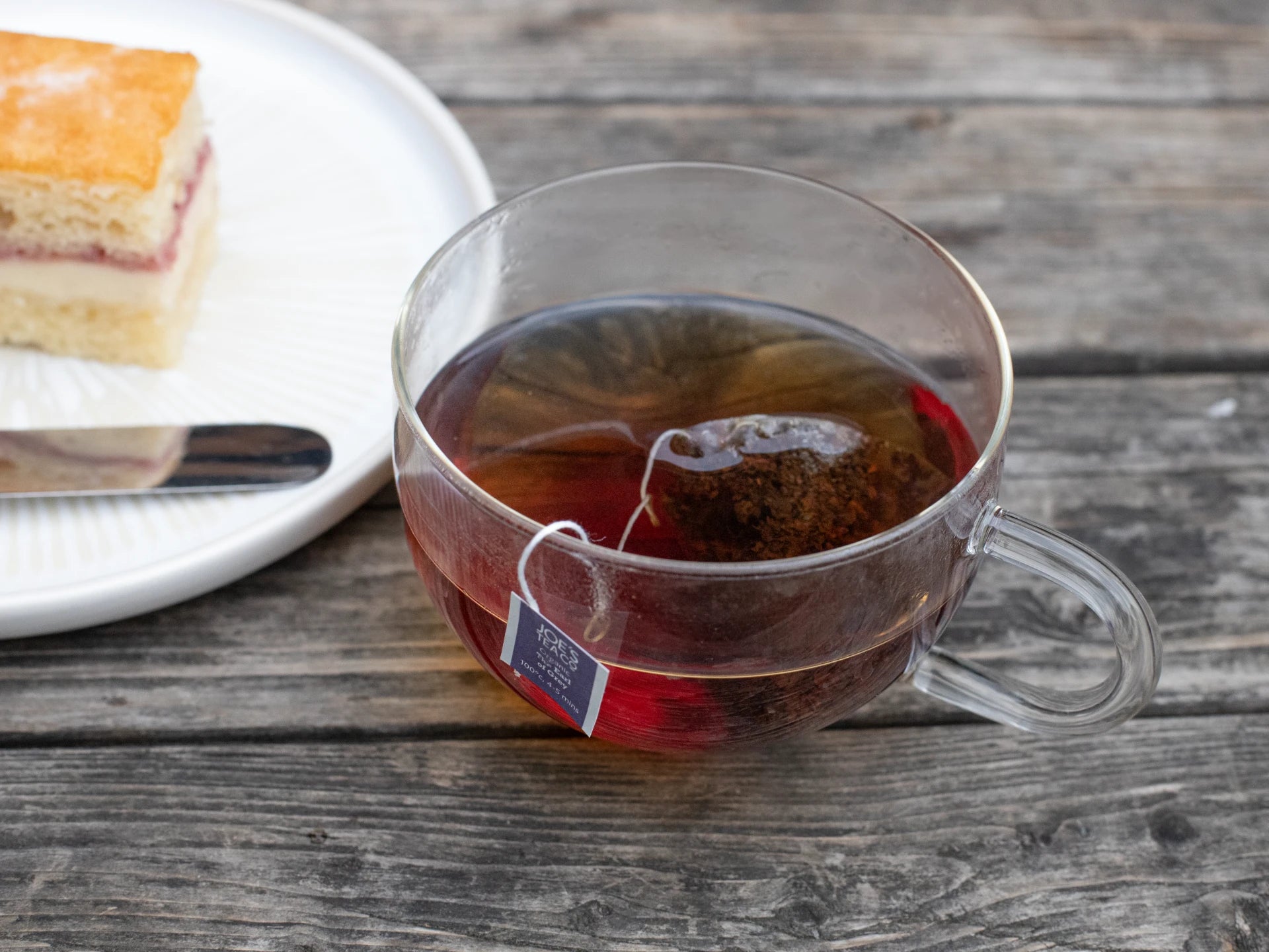 Clear glass mug with Joe's Tea co  tea and a tea bag on a wooden table with a plate of cake.