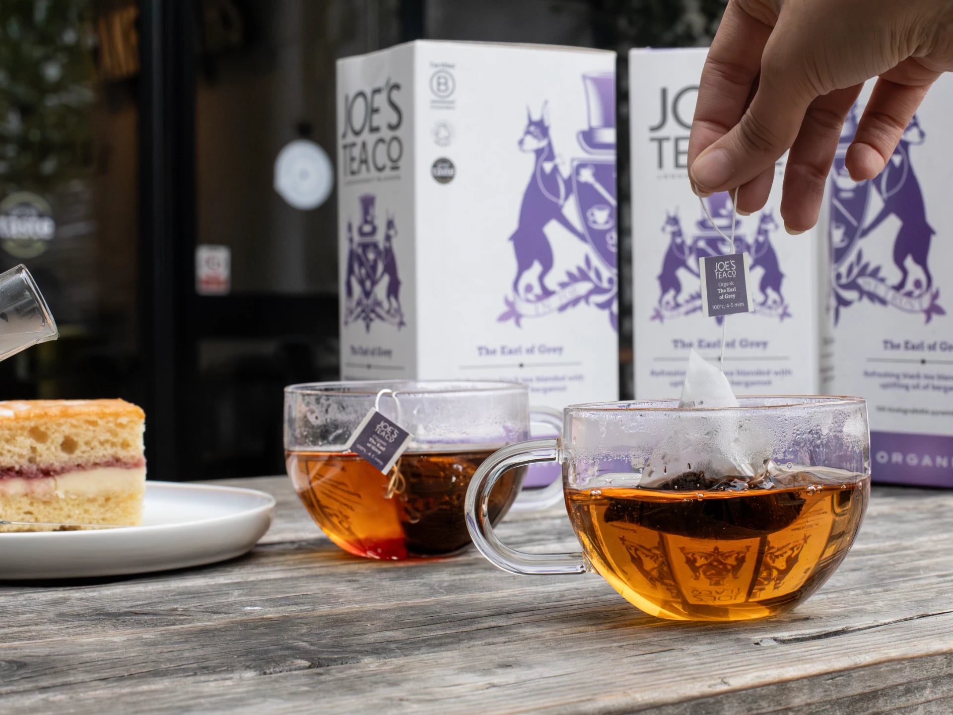 Two cups of tea with a hand reaching for a box of Joe's Tea Co. on a wooden table.