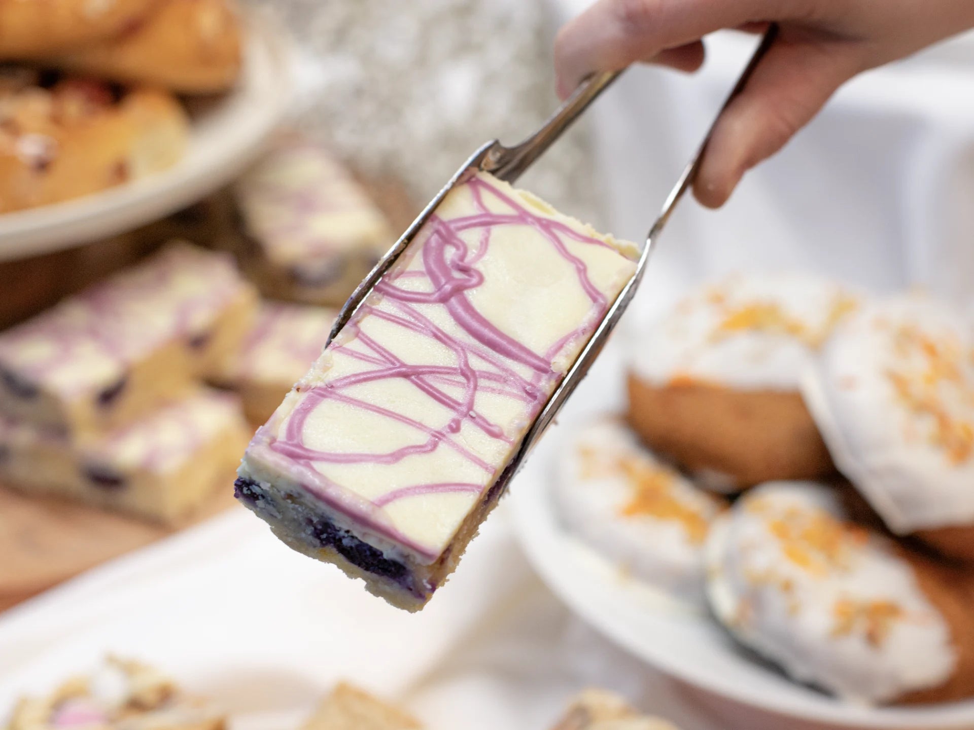 Slice of White Chocolate Blueberry Blondie with purple drizzle held by a spatula over other desserts.